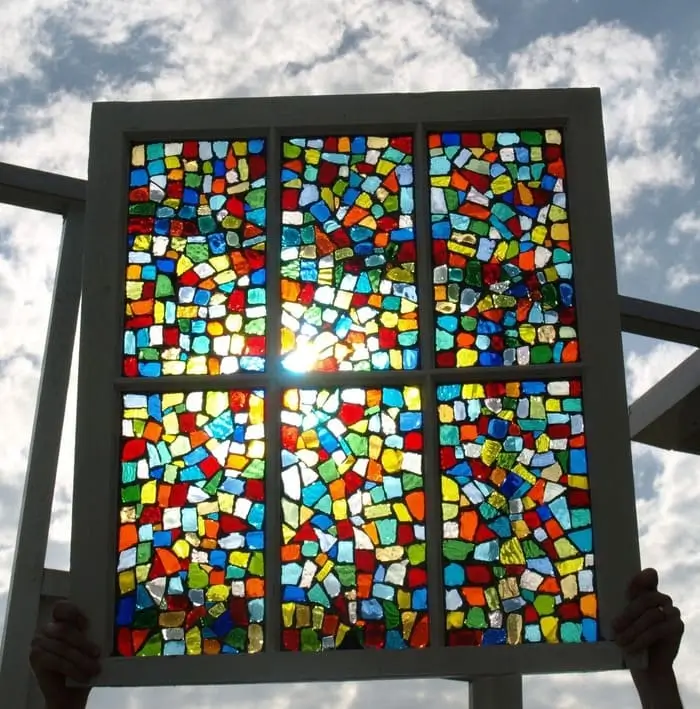 A person holding a multi-pane window frame filled with a colorful mosaic of small stained glass pieces against a cloudy sky